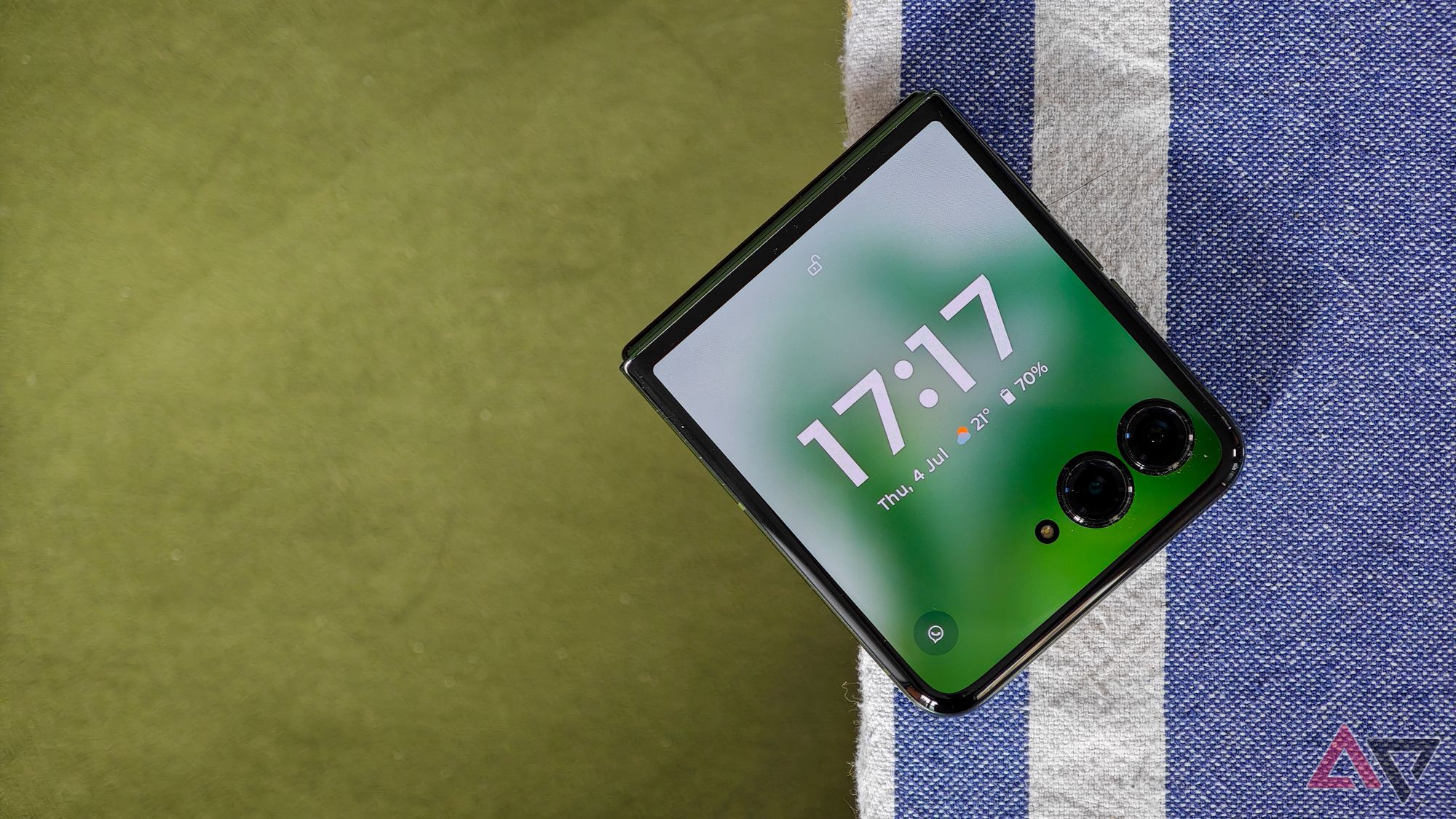 Motorola Razr+ showing the time on the cover screen, resting on a blue tablecloth above a green velvet background